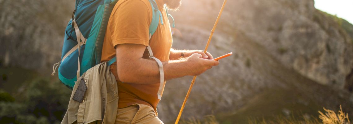 Auf dem Bild ist ein wandernder Mann in der Natur abgebildet, der einen Wanderstock hält und einen Rucksack auf dem Rücken trägt. 